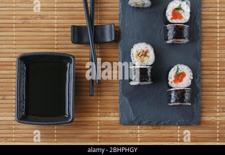 Des petits pains à sushis au saumon, au thon, à l'avocat, au riz aux algues et des baguettes sur un tableau sombre allongé sur un tapis de bambou. Japonais, Banque D'Images