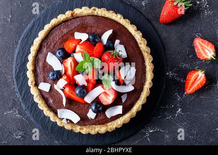 Tarte française au chocolat noir soyeux, décorée de fraises et de bleuets, de chips de coco sur le dessus, servie sur fond de béton foncé Banque D'Images