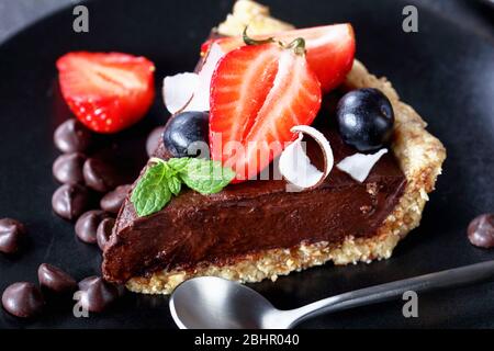 Tarte au chocolat végétarien avec fraises fraîches, bleuets, chips de noix de coco sur le dessus avec croûte sans cuisson d'avoine roulée, dattes et noix se Banque D'Images