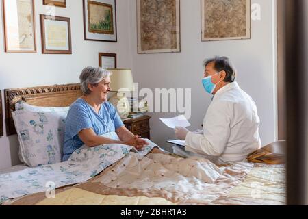 Un médecin en manteau blanc et un masque de protection pour le visage faisant une visite à domicile chez une femme âgée. Banque D'Images