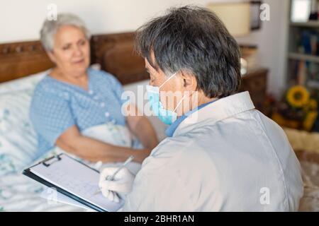 Un médecin en manteau blanc et un masque de protection pour le visage faisant une visite à domicile chez une femme âgée. Banque D'Images