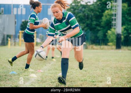 Trois femmes portant des maillots de rugby bleus, blancs et verts sur un terrain d'entraînement, une courir avec un ballon de rugby. Banque D'Images