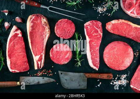 Morceaux de variété de viande, tiré de dessus sur un fond noir avec sel, poivre, romarin et couteaux, une couche plate