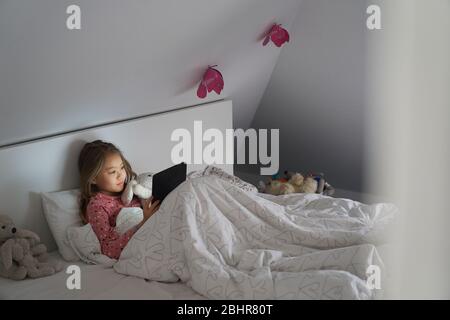 La vie à la maison, un matin scolaire pendant le verrouillage. Une fille couchée au lit à l'aide d'une tablette numérique. Banque D'Images