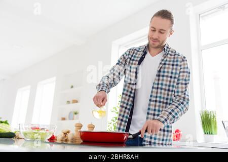 Photo de positif beau homme cuisinier délicieux repas de souper verser de l'huile de tournesol sur la poêle porter décontracté carreaux plat chemise debout dans la cuisine Banque D'Images
