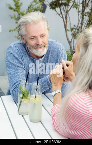 Un couple assis à une table dans un jardin avec des boissons en bouteilles, regardant l'un l'autre et tenant les mains. Banque D'Images