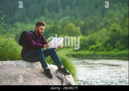 Un homme touristique barbu, sur le rivage d'une rivière de montagne, étudie une carte. Banque D'Images