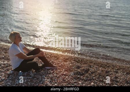 Femme mûre assise après le jogging sur la plage au coucher du soleil. Course en soirée Banque D'Images
