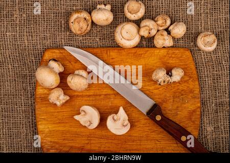 Moitiés de jeunes champignons coupés et un couteau avec poignée en bois sur un panneau de découpe en bois et champignons dispersés sur un fond de gros homespun fa Banque D'Images