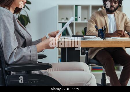 Employé handicapé en fauteuil roulant avec des papiers et recruteur africain américain à la table au bureau Banque D'Images