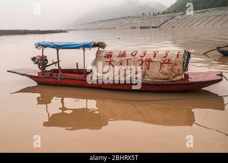 Fengdu, Chine - 8 mai 2010 : gros plan de la petite boucle couverte et son reflet dans l'eau marron de la rivière Yangtze, avec des rives dans le brouillard et le mandarin rouge c Banque D'Images