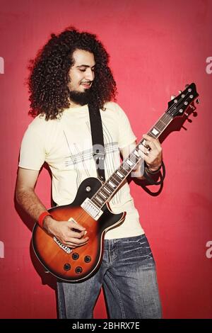 Gros plan d'un bel passionné expressif jeune homme musicien de rock brunette cool avec des cheveux longs jouant de l'électro guitare contre fond rouge Banque D'Images
