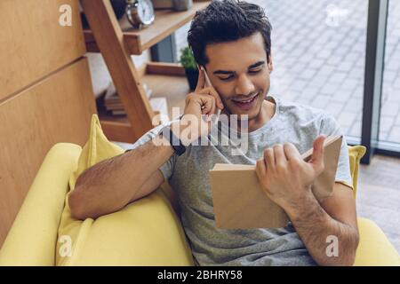 Vue sur l'homme avec livre souriant et parler sur smartphone sur canapé dans le salon Banque D'Images