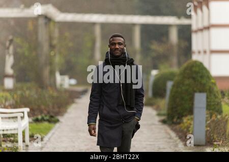 Jeune homme noir bien habillé portant des écouteurs et un manteau noir posant dans le parc d'automne. Banque D'Images