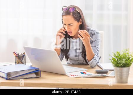 Bouleversée femme d'affaires hurlant dans le téléphone mobile gestuing, tout en étant assis devant son ordinateur au bureau. Banque D'Images