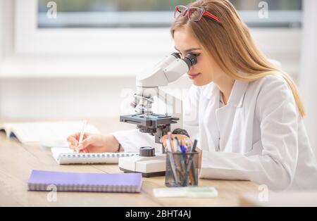 Belle jeune étudiante chercheuse regardant dans un microscope, écrivant des notes. Banque D'Images