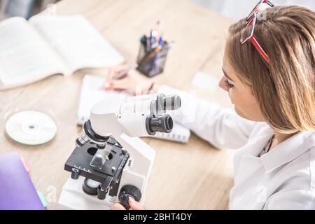 Technicien de laboratoire féminin qui écrit des observations à partir du travail au microscope. Banque D'Images