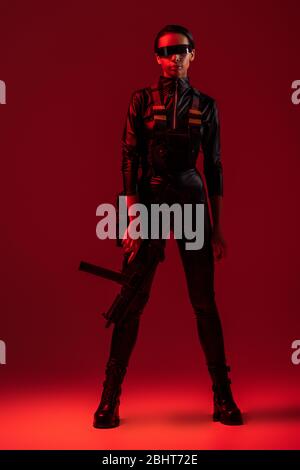 une femme afro-américaine futuriste en lunettes avec fusil d'assaut sur fond rouge Banque D'Images