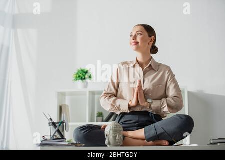 Une femme d'affaires heureuse méditant dans lotus pose avec un geste namaste sur le lieu de travail avec la tête de Bouddha et le bâton d'encens Banque D'Images