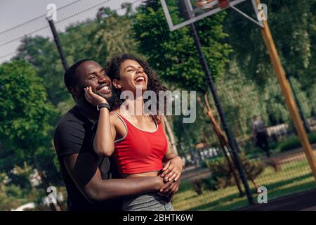 Activité en plein air. Couple africain debout sur un terrain de basket-ball embrassant rire heureux Banque D'Images