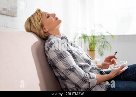 vue latérale d'une femme mûre avec des yeux fermés assis sur un canapé et tenant conjoint avec la marijuana légale Banque D'Images