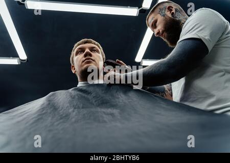 Coupe de cheveux avec rasoir électrique. Le tatttooed Barber fait la coupe de cheveux pour le client au salon de coiffure en utilisant la tondeuse à cheveux. Homme coiffure avec électrique Banque D'Images