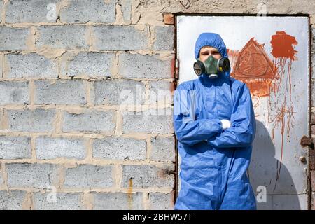 homme dans des combinaisons et un masque bleus de protection debout près d'un mur de briques, bâtiment abandonné. Banque D'Images