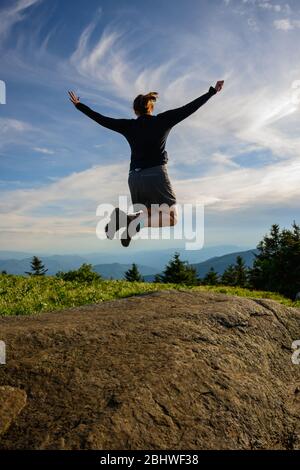 La femme lève les bras dans Leap of Joy sur la crête bleue de montagne surplombant Banque D'Images