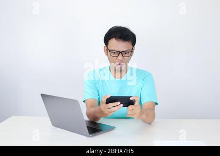 Un jeune homme asiatique en colère se fait fou sur son smartphone lorsqu'il joue au travail. Homme indonésien portant une chemise bleue. Banque D'Images