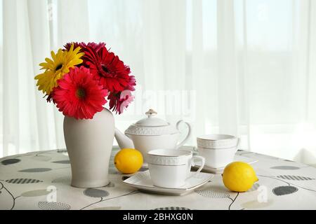 Gerbera coloré dans un vase avec théière, tasses et citrons sur table sur le fond des rideaux Banque D'Images