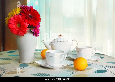 Gerbera coloré dans un vase avec théière, tasses et citrons sur table sur le fond des rideaux Banque D'Images