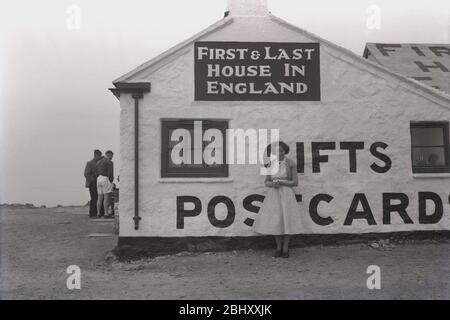 Années 1950, historique, jeune femme, de la vingtaine, dans une robe de l'époque posant pour sa photo se tenant à l'extérieur de la célèbre 'première et dernière maison' à Sennen, Lands End, Cornwall,. Le vieux cottage en pierre blanchi à blanc vendait des rafraîchissements et des souvenirs et situé sur la côte déchiquetée à l'ouest de l'Angleterre fonctionne toujours comme tel aujourd'hui. Banque D'Images