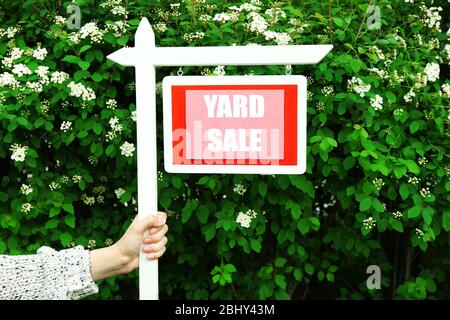 Panneau de vente de Yard en bois dans la main femelle sur le buisson vert et les fleurs fond Banque D'Images