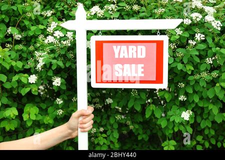 Panneau de vente de Yard en bois dans la main femelle sur le buisson vert et les fleurs fond Banque D'Images