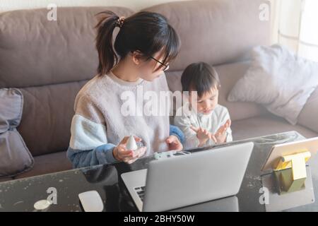 Jeune mère asiatique travaillant sur un ordinateur portable moderne de la maison avec son enfant regardant la bande dessinée sur un comprimé pendant le verrouillage de la pandémie de coronavirus Banque D'Images