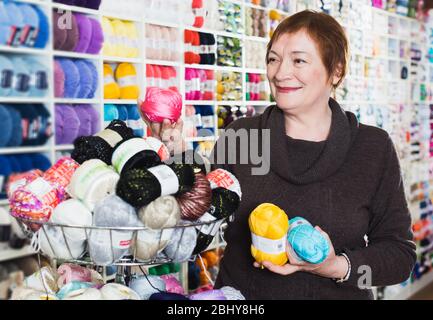 Femme âgée positive tenant des fils colorés à tricoter sur le fond des étagères du magasin Banque D'Images