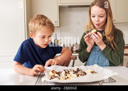 Frère et sœur mangeant des petits pains à la cannelle fraîchement cuits dans la cuisine. Banque D'Images