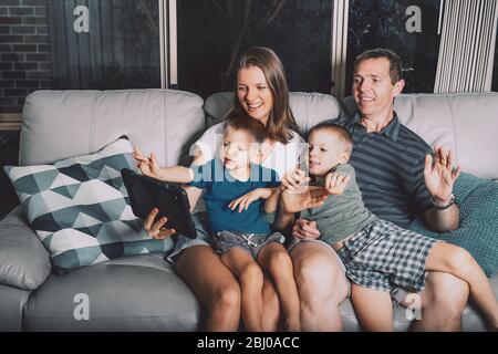 Famille de parents et deux petits fils communiquant avec des parents par appel vidéo sur tablette électronique tout en se reposant sur canapé dans la maison. Distance sociale pendant le verrouillage de la quarantaine Banque D'Images