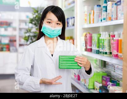 Diligent sympathique souriante femme coréenne pharmacien dans le masque facial protecteur posant avec des médicaments dans la pharmacie moderne Banque D'Images