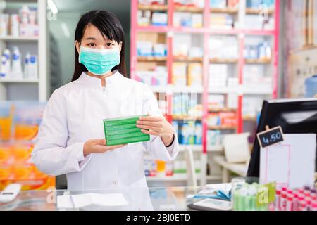 Diligent souriant agréable femme coréenne pharmacien dans le masque facial protecteur posant avec des médicaments en pharmacie moderne Banque D'Images