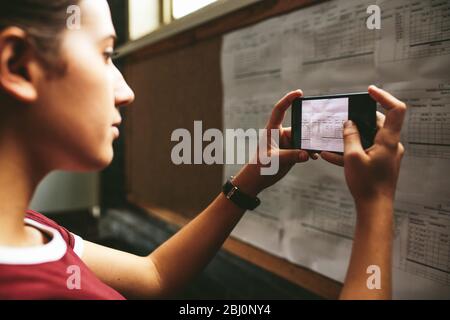 Une étudiante en femme qui parle une photo du conseil d'administration du lycée. Fille prenant une photo de l'horaire d'examen avec son téléphone portable à l'école. Banque D'Images