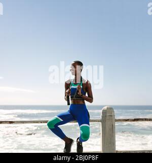 Heureuse femme africaine dans des vêtements de sport assis sur la rampe sur la route de bord de mer tenant une corde à sauter. Femme prenant une pause après avoir fait de l'exercice à l'extérieur. Banque D'Images