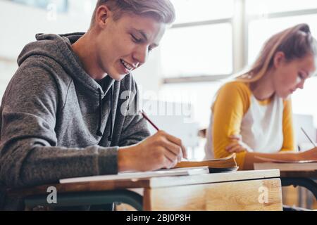 Adolescent assis dans la classe de lycée écriture dans le livre. L'étudiant masculin prend des notes pendant la conférence en classe universitaire. Banque D'Images