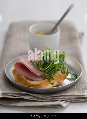 Sandwich ouvert de pain croustillant rouleau rustique, avec une tranche épaisse de jambon de qualité et une garniture de feuille de salade de roquette, avec une petite casserole de moutarde de Dijon - Banque D'Images