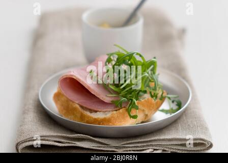 Sandwich ouvert de pain croustillant rouleau rustique, avec une tranche épaisse de jambon de qualité et une garniture de feuille de salade de roquette, avec une petite casserole de moutarde de Dijon - Banque D'Images