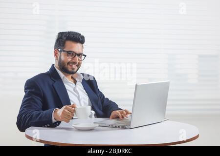 Jeune homme assis dans son bureau tout en travaillant sur l'ordinateur portable pendant que vous buvez du café. Banque D'Images