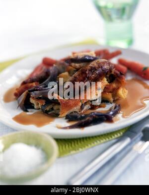 Plat britannique classique de Toad in the Hole, pâte à pâte cuite avec saucisses, servi avec sauce à l'oignon rouge et carottes bouillies - Banque D'Images