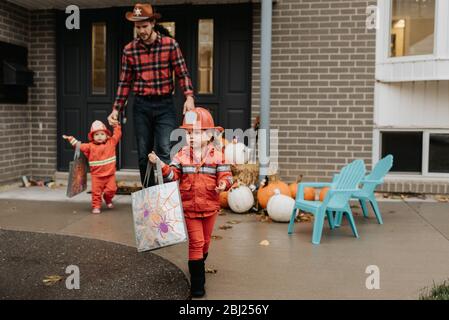 Deux enfants vêtus de pompiers marchant loin de la porte d'entrée d'une maison décorée pour Halloween avec leur père. Banque D'Images