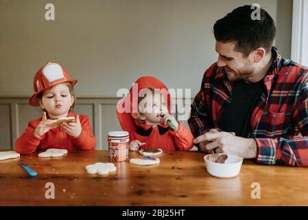 Deux enfants vêtus de pompiers et leur père à une table décorant des biscuits avec des chocolats et des saupoudrés. Banque D'Images