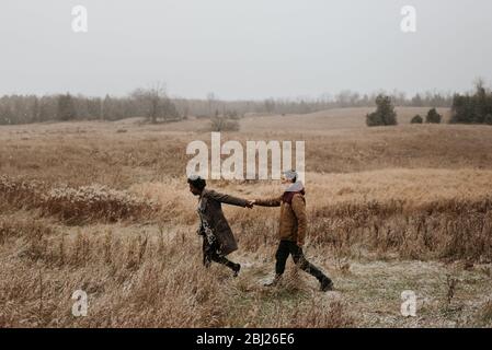 Une femme noire et un homme blanc qui tient les mains et marche à travers les herbes dans un paysage rural de wintry. Banque D'Images